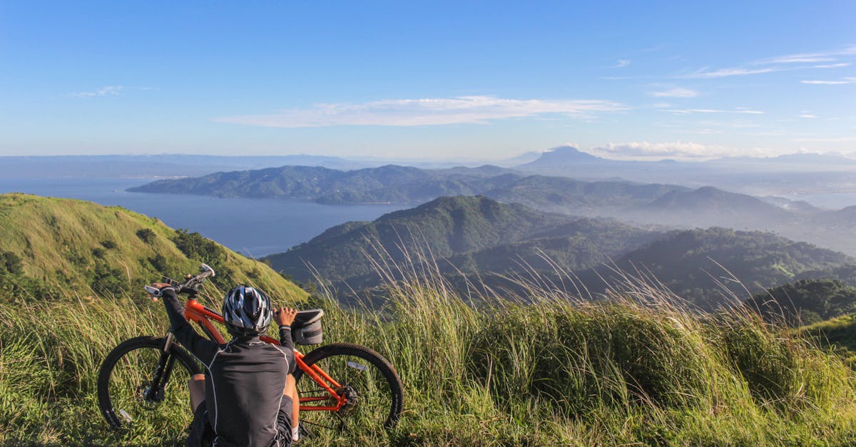 A mountain biker resting by their bike, enjoying a scenic view of lush hills and distant water under a bright blue sky.